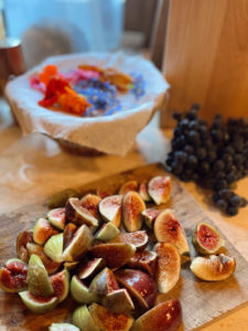 Kitchen counter: A cutting board with figs quartered, dark purple grapes and a bowl covered in muslin and flowers in the background