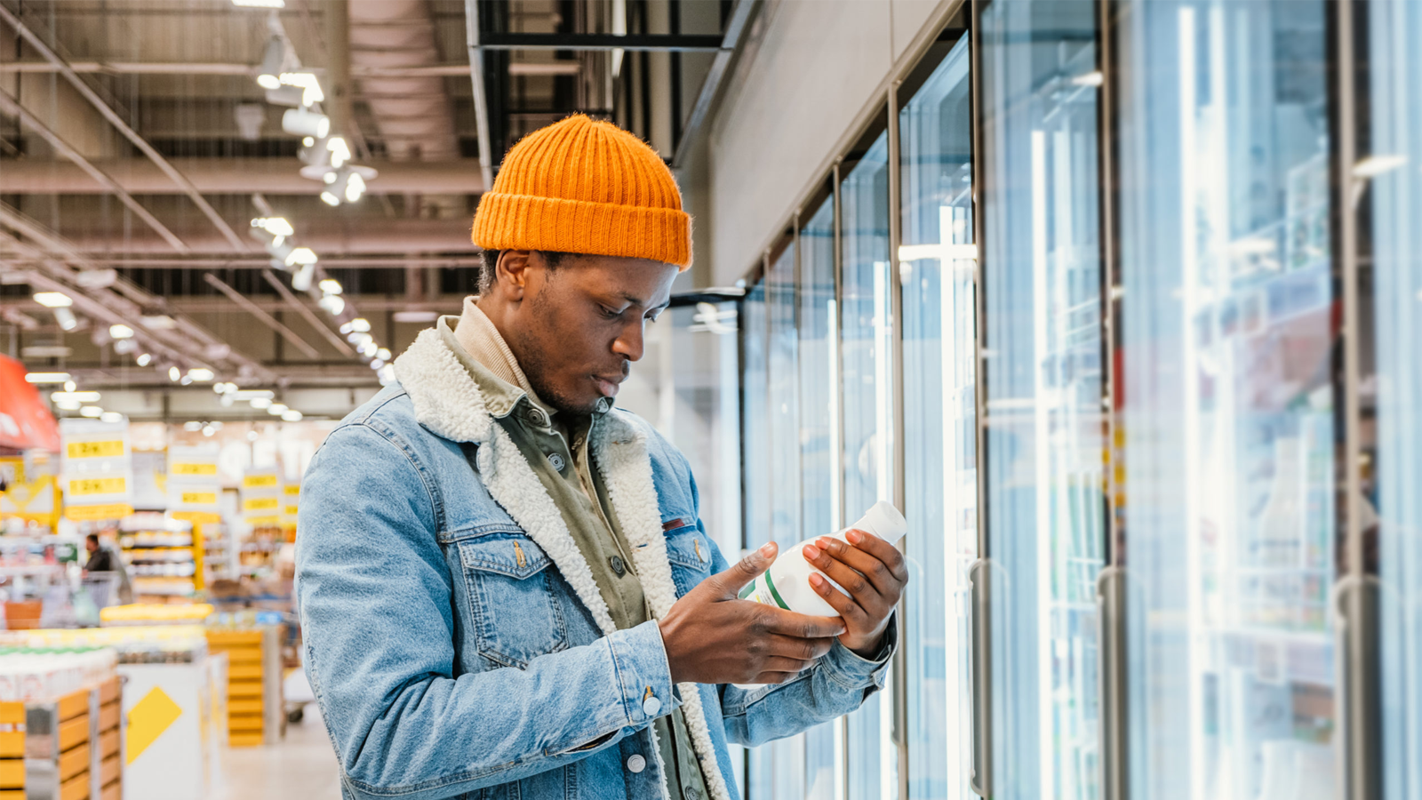 Positive African-American guy in denim jacket and orange hat takes milk bottle from fridge case in modern supermarket side view