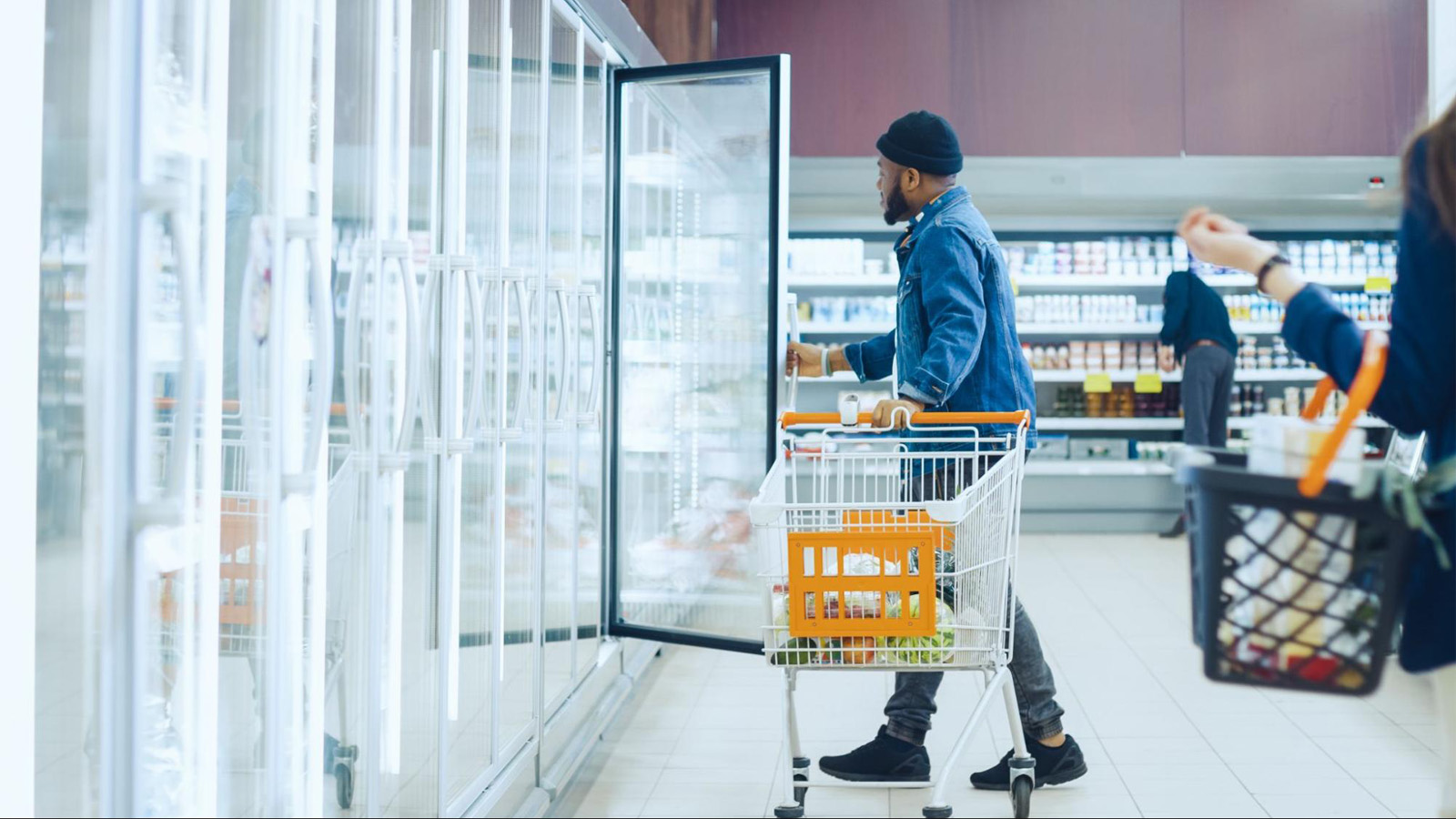 Man standing with a shopping cart opening a refrigerator door in a grocery store
