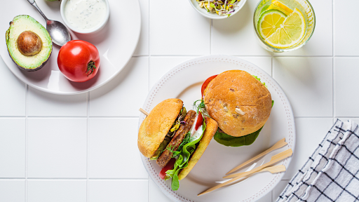 A pair of juicy, plant-based burgers ready-to-eat on a white plate, topped with lettuce, tomato, avocado and tzaziki. There's a glass of water flavored with citrus slices beside the burgers.