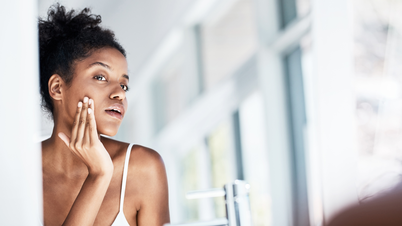 African American woman with curly hair tied up looking in the mirror doing skincare