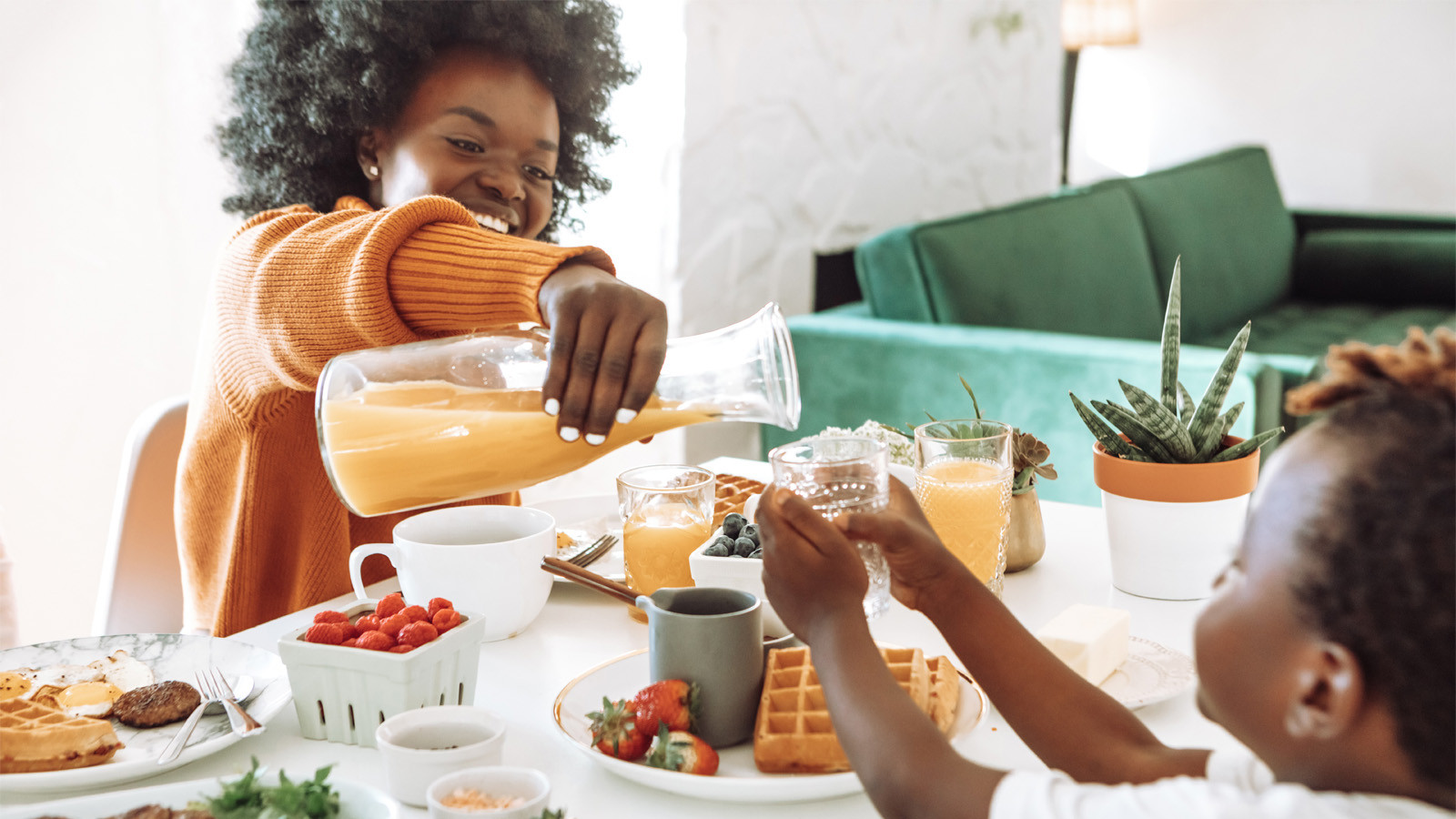 Woman of color pouring orange juice from a carafe to a child on the breakfast table