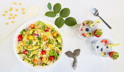 Aerial view of a white table with a mixed grain and vegetable dish on the left and bowls of chia seeds with fruit on the right. Chickpeas, decorative greenery and grains in leaf shapes surround the dishes of food.