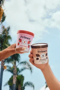 Pints of Fronen strawberry and Fronen chocolate dairy free frozen dessert are held up against a tropical sky.