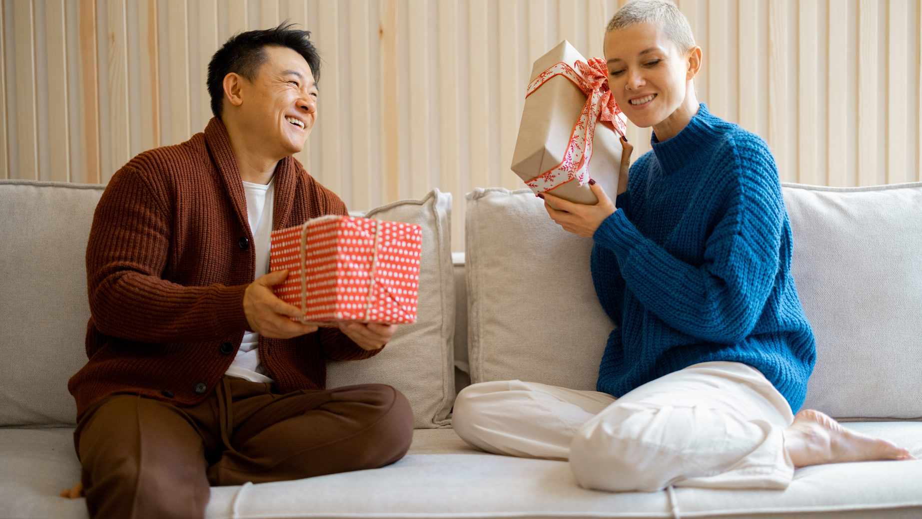 Asian American man and Caucasian woman sitting on a couch both holding in their hands wrapped holiday gifts