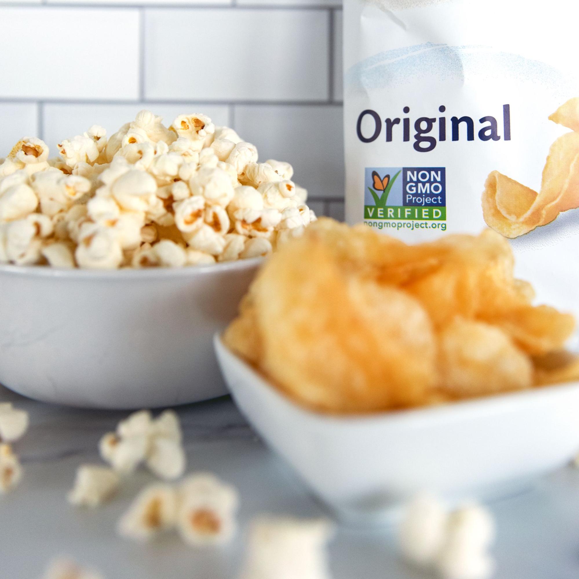 A bowl of popcorn and a bowl of potato chips sitting in front of a bag of chips with the Non-GMO Project Verified mark on package