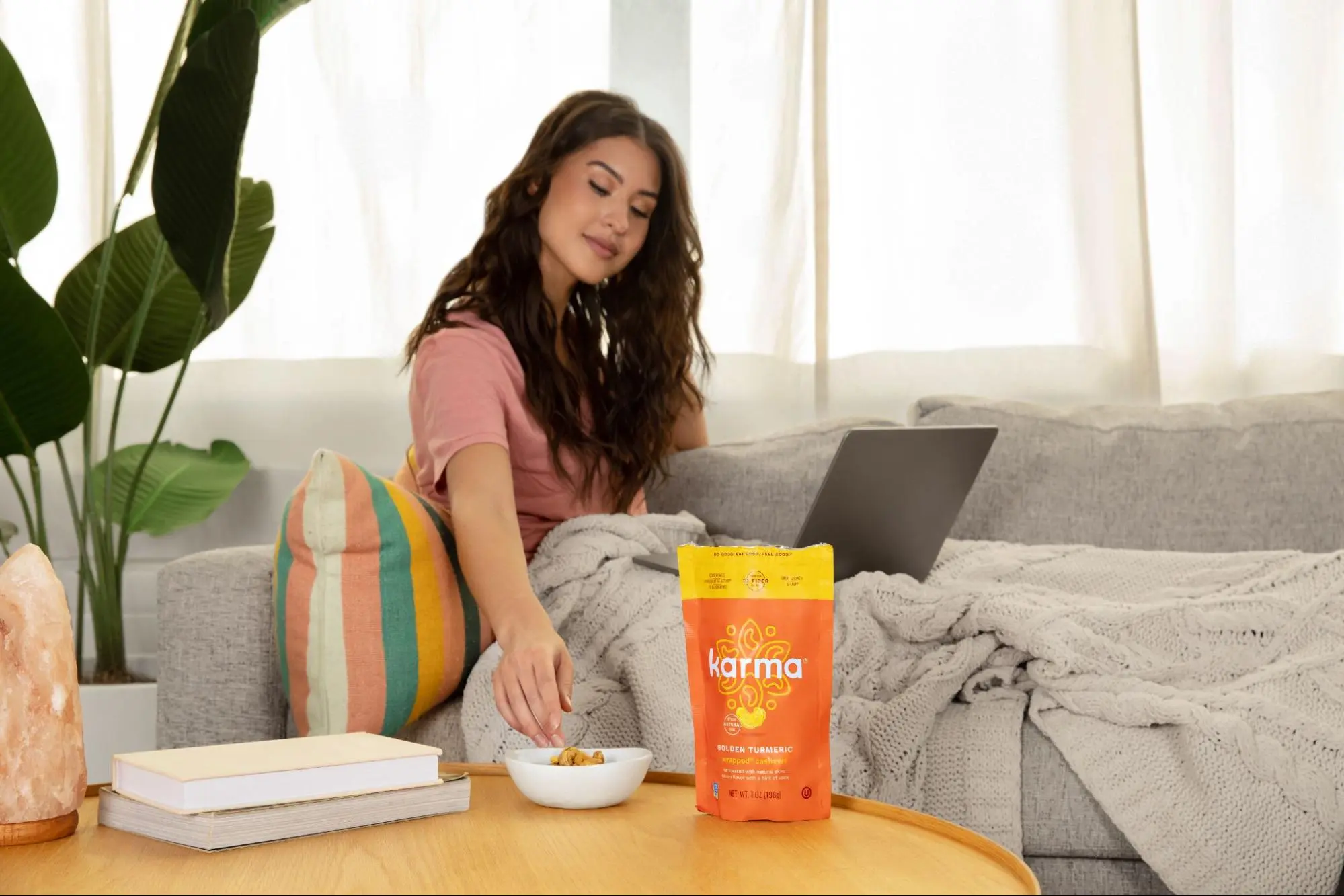 A woman relaxes on a couch, using her laptop and eating Karma Nut cashews from a bowl.
