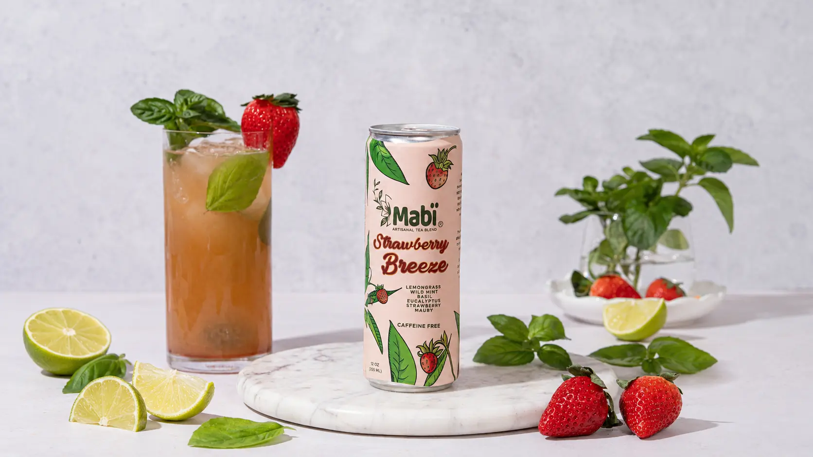 A can of Strawberry Breeze Mabi Artisanal Tea sits on a marble tray, surrounded by bright red strawberries, deep green basil and cool lime wedges.