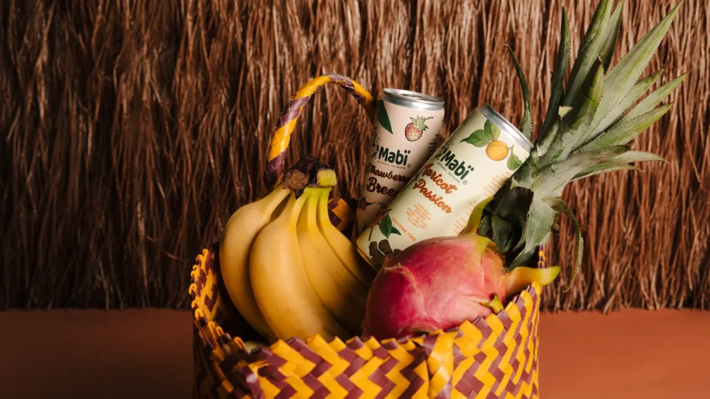 A tote bag filled with tropical fruit and Mabi Artisanal tea sits in front of a coconut fiber backdrop.