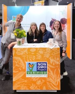 Non-GMO Project four staff members standing and smiling behind Natural Products Expo West tradeshow booth.