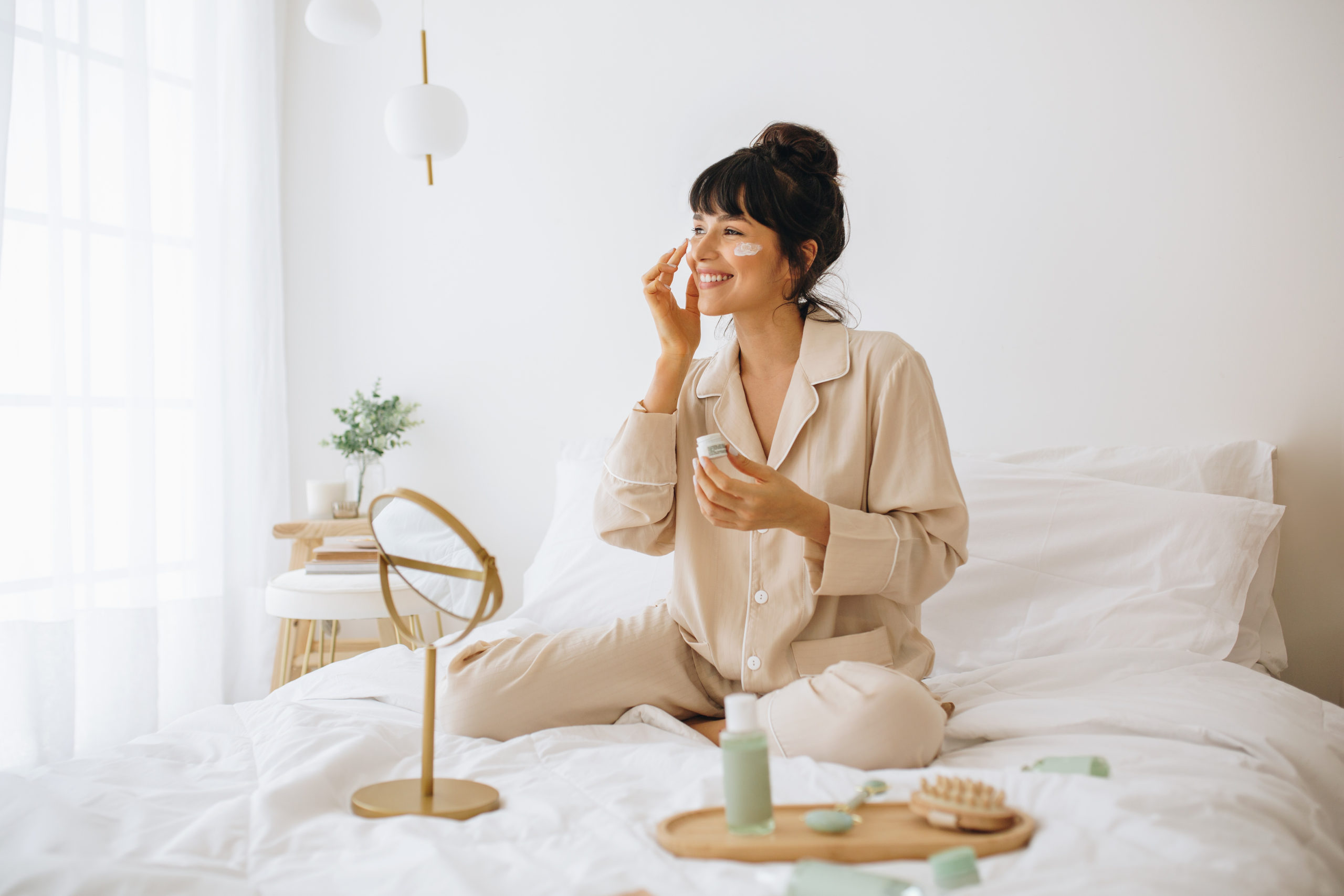 Happy woman doing routine skin care at home with beauty products. Woman sitting on bed at home and applying face cream.