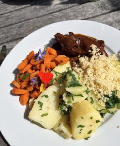 A white dinner plate on an outdoor table. The meal is couscous, stewed meat, potatoes and carrots, garnished with parsley and flowers.
