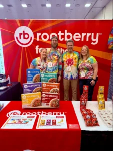 Four people wearing tie-dye t-shirts stand behind a table displaying of rootberry products and information, smiling and engaging with attendees.