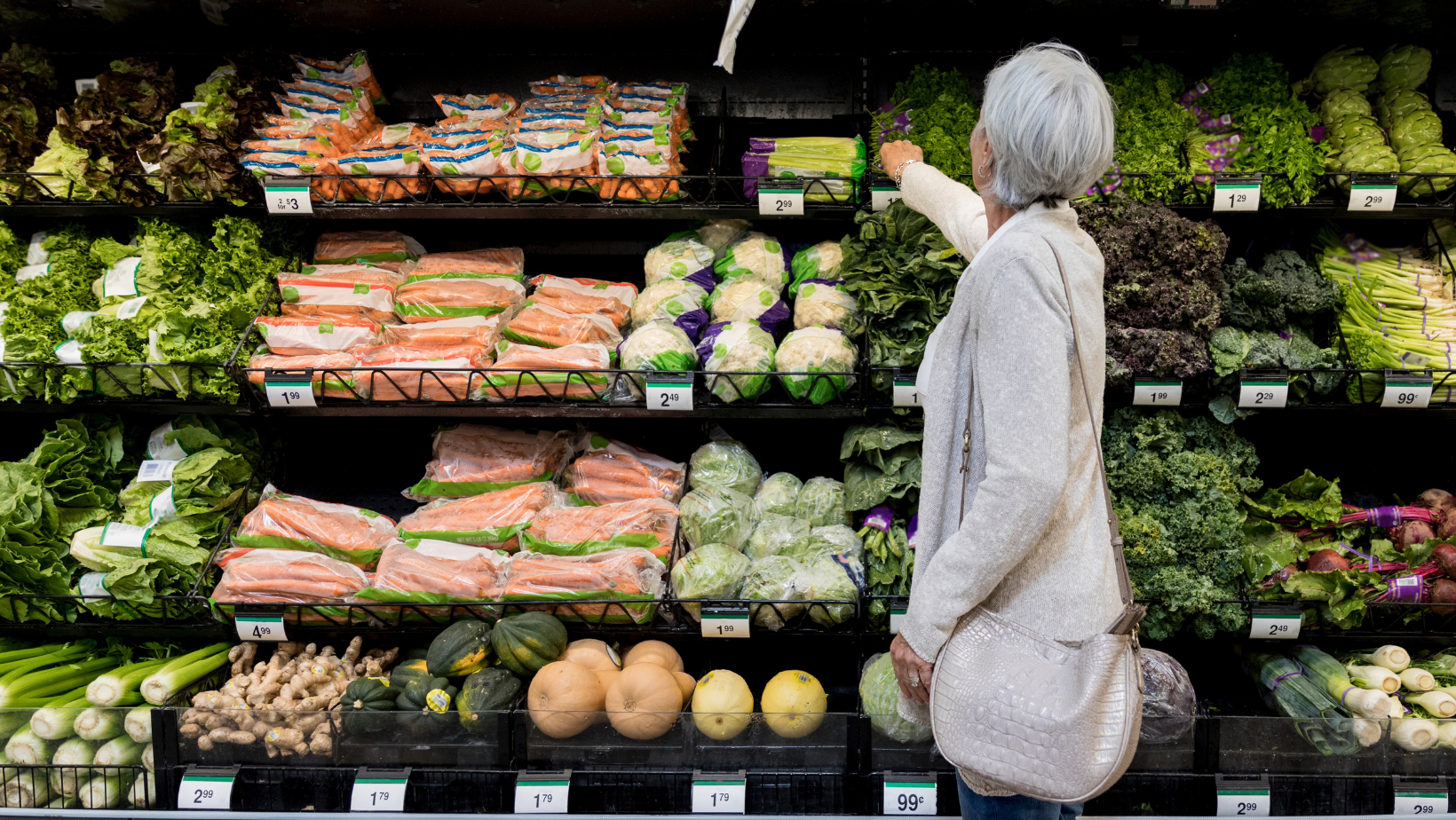 Older white woman wearing a gray cardigan is standing in the vegetable section of a grocery store